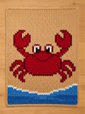 Chunky knit blanket featuring a large red crab design on a sandy beige background with blue and white ocean waves at the bottom, laid flat on a wooden floor