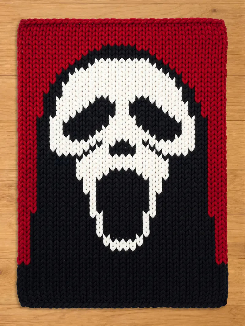 A knitted rectangular throw blanket featuring a white skull design against a black background. The top and sides are framed with a red border. The skull has a wide open mouth.