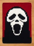 A knitted rectangular throw blanket featuring a white skull design against a black background. The top and sides are framed with a red border. The skull has a wide open mouth.