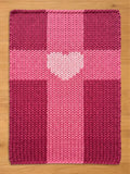 A knitted blanket in shades of pink and magenta, featuring a heart-shaped design in the center. The blanket has a textured appearance, with a cross-like pattern. The background is a light brown wooden surface.