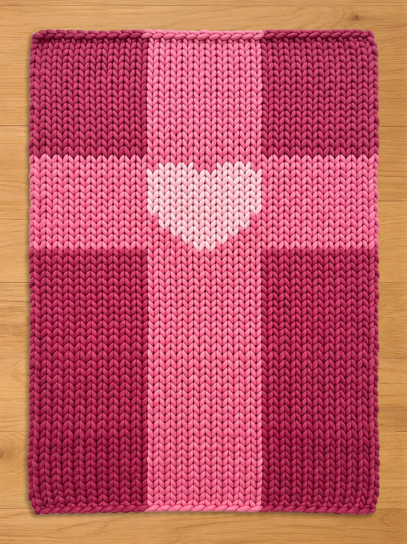 A knitted blanket in shades of pink and magenta, featuring a heart-shaped design in the center. The blanket has a textured appearance, with a cross-like pattern. The background is a light brown wooden surface.