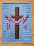 A knitted blue chunky blanket with a brown cross at the center, adorned with a purple and pink garland. The cross is a darker shade of brown, and the garland adds a decorative touch to the religious-themed design.