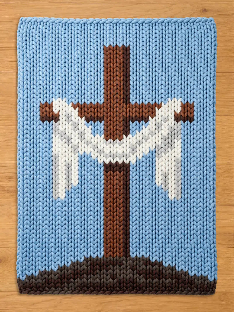 A knitted blanket featuring a brown cross with white accents against a light blue background. The cross is set on a brown and gray base, with a light blue border. The blanket is made of a soft, textured material.