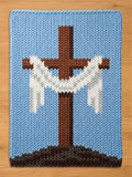 A knitted blanket featuring a brown cross with white accents against a light blue background. The cross is set on a brown and gray base, with a light blue border. The blanket is made of a soft, textured material.