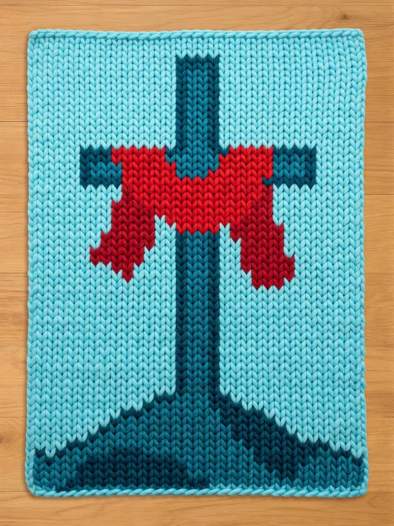 A knitted chunky blanket featuring a cross design. The background is light blue, with a darker blue cross and a red accent. The piece is made of thick yarn, giving it a textured appearance. The design is simple and geometric.