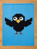 A light blue chunky knit blanket featuring a cartoon black crow or bird design in the center. The crow has big white eyes, a yellow beak, and brown feet, knitted with thick, textured yarn. Placed flat on a light wood floor.