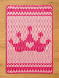 A knitted pink and light pink rectangular chunky blanket with a crown design. The crown is a darker shade of pink and is centered on the mat. The blanket has a border and stripes in a darker pink.