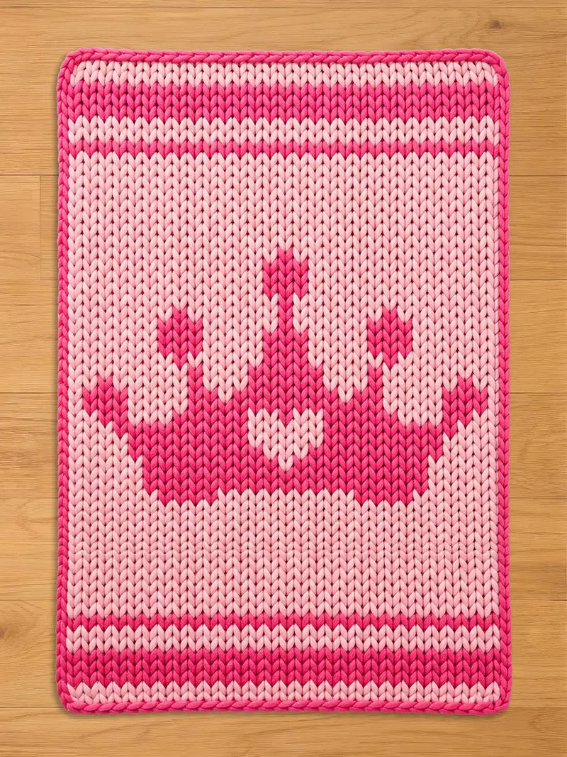 A knitted pink and light pink rectangular chunky blanket with a crown design. The crown is a darker shade of pink and is centered on the mat. The blanket has a border and stripes in a darker pink.