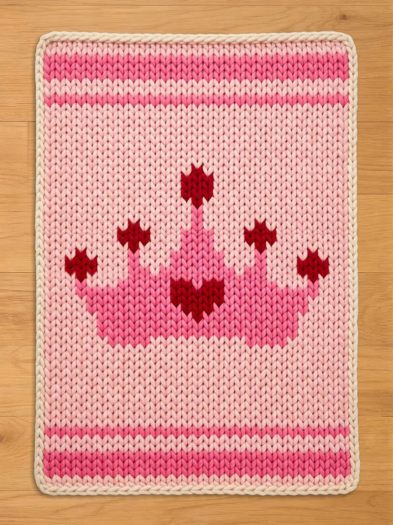 Pink chunky knit blanket featuring a princess crown and heart design. This handmade-style knit throw blanket includes hot pink borders, a cream trim, and a red heart crown motif, perfect for a nursery or kids' bedroom.