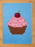 Light blue chunky knit blanket featuring a woven design of a cupcake with pink frosting, brown liner, sprinkles, and a red cherry on top, displayed on a wooden floor.