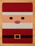 A top-down view of a thick, chunky knit blanket featuring a stylized Santa Claus face and suit design lying on a light wood floor. The blanket has a red top and bottom, a white trim and beard section, a peach-colored face with black knit eyes and pink lips, and a black belt with a yellow buckle at the bottom.