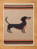 A knitted rectangular chunky blanket featuring a dachshund dog design. The dog is black and peach, set against a beige background. The blanket has a cream border and stripes in black and orange.
