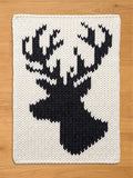 A rectangular, off-white chunky knit blanket displaying a prominent, contrasting black silhouette of a deer head with full antlers in the center. The blanket is resting flat on a light wood floor.