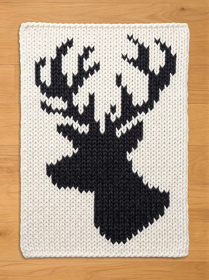 A rectangular, off-white chunky knit blanket displaying a prominent, contrasting black silhouette of a deer head with full antlers in the center. The blanket is resting flat on a light wood floor.