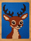 A knitted  chunky blanket featuring a cartoon deer with brown antlers, a brown body, and a white chest. The deer has large black eyes and a black nose. The background is a solid blue color.