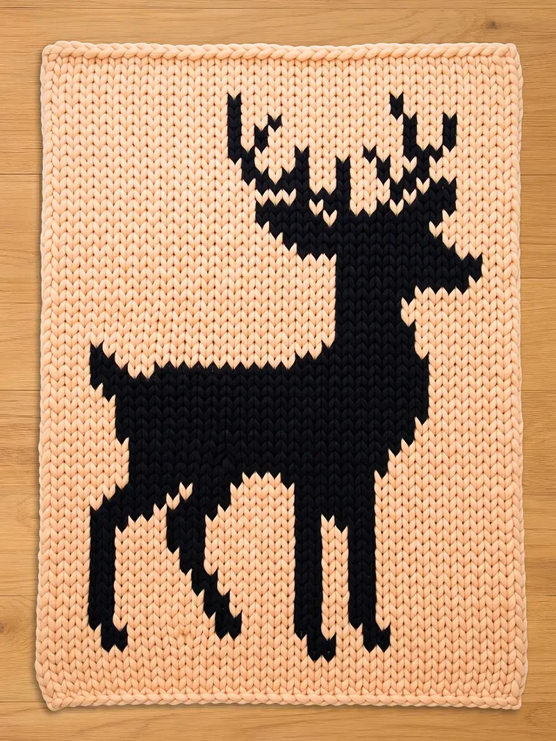 A knitted chunky blanket featuring a black deer silhouette against a light peach background. The deer design is a simple, geometric shape. The blanket is made of thick yarn, giving it a textured appearance.