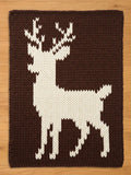 A chunky knit blanket in dark brown featuring a large, white knit stag deer silhouette with prominent antlers, displayed flat on a wooden surface.