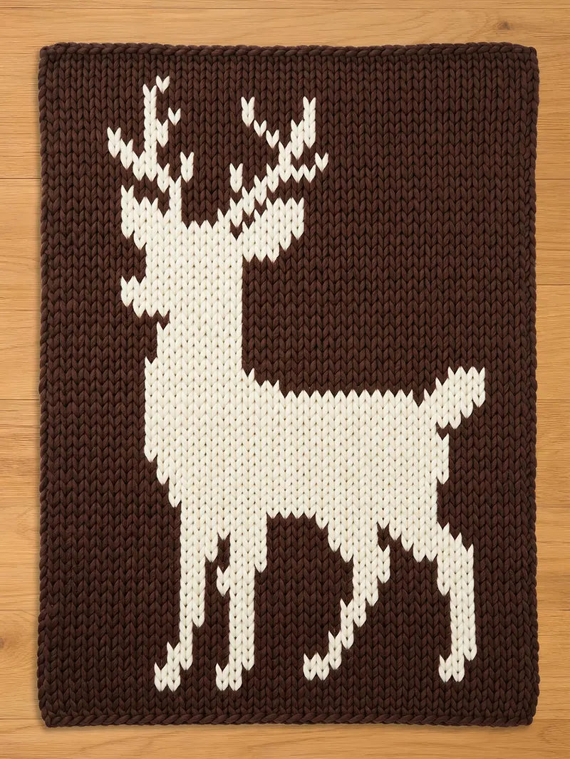 A chunky knit blanket in dark brown featuring a large, white knit stag deer silhouette with prominent antlers, displayed flat on a wooden surface.