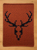 A rectangular, rust-colored knitted chunky blanket featuring a black deer head design. The blanket has a thick, textured appearance, with the deer silhouette prominently displayed in the center. The background is a warm, earthy tone.