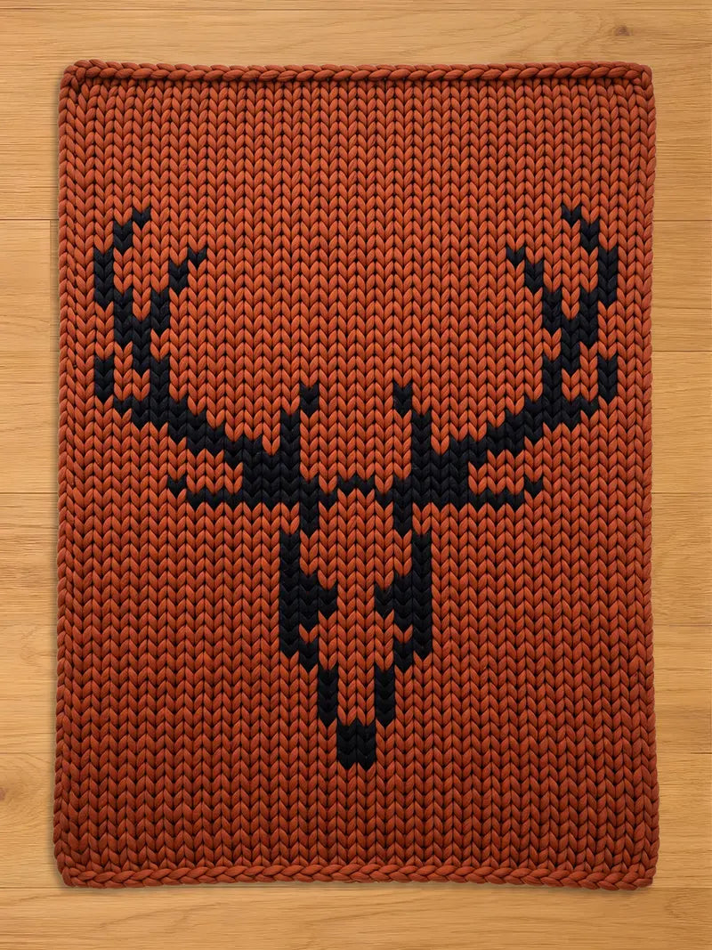 A rectangular, rust-colored knitted chunky blanket featuring a black deer head design. The blanket has a thick, textured appearance, with the deer silhouette prominently displayed in the center. The background is a warm, earthy tone.