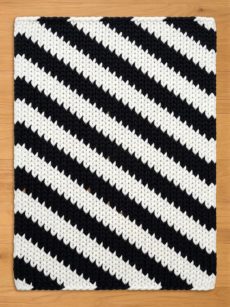 A black and white knitted chunky blanket with a bold diagonal stripe pattern. The blanket is rectangular and features thick, textured stripes. The contrasting colors create a striking visual effect, suitable for home decor.