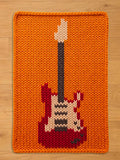 An orange knitted rectangular chunky blanket featuring a red, white, black, and gray electric guitar design. The guitar has a black neck and a white headstock. The blanket  is made of thick yarn.