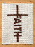 White chunky knit blanket featuring the word "FAITH" knitted vertically in brown to form a cross design, displayed on a wooden floor.