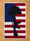 A knitted rectangular chunky knit blanket featuring a black silhouette of a soldier's memorial against a backdrop of the American flag. The flag design includes blue, red, and white stripes and a white border.