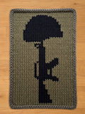 A chunky knit blanket featuring a black silhouette of a helmet and rifle, set against a khaki green background. The blanket has a gray border and is made of a thick, textured yarn. The design is a tribute to fallen soldiers.