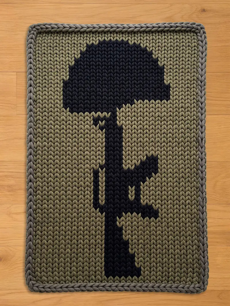 A chunky knit blanket featuring a black silhouette of a helmet and rifle, set against a khaki green background. The blanket has a gray border and is made of a thick, textured yarn. The design is a tribute to fallen soldiers.