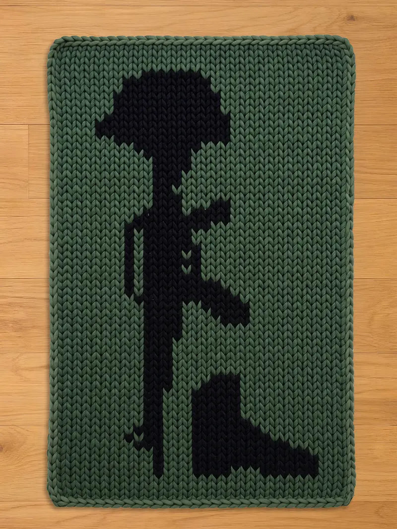 A knitted green and black chunky blanket featuring a black silhouette of a soldier's memorial. The design includes a helmet, rifle, and boots, all in a dark color against a green background. The blanket has a rectangular shape.