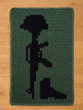 A knitted green and black chunky blanket featuring a black silhouette of a soldier's memorial. The design includes a helmet, rifle, and boots, all in a dark color against a green background. The blanket has a rectangular shape.