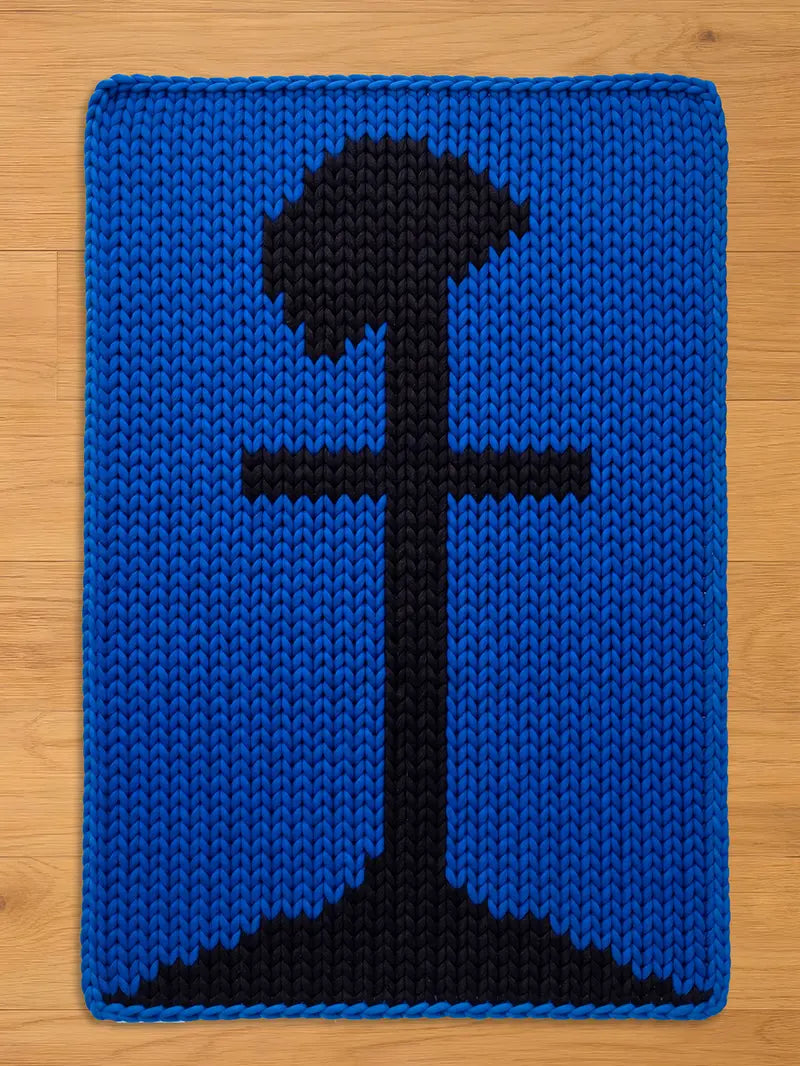 A knitted blue rectangular chunky blanket with a black graphic design. The design features a stylized black tree or abstract shape in the center. The blanket is made of thick, textured yarn.