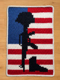 A knitted blanket featuring the American flag with a black silhouette of a rifle, helmet, and boots, symbolizing fallen soldiers. The blanket has a blue field with white stars, red and white stripes, and a gray border.