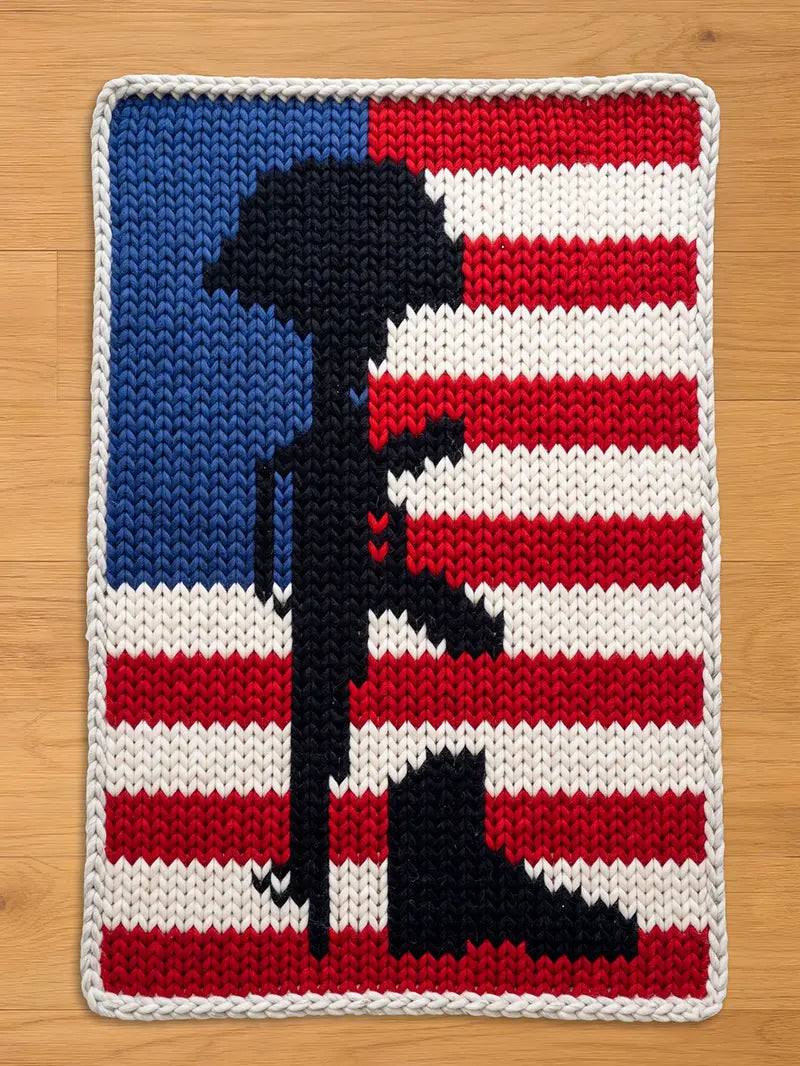 A knitted blanket featuring the American flag with a black silhouette of a rifle, helmet, and boots, symbolizing fallen soldiers. The blanket has a blue field with white stars, red and white stripes, and a gray border.