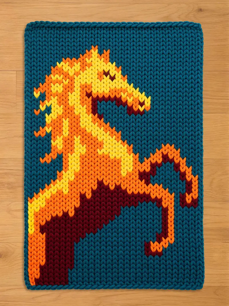 A knitted chunky blanket featuring a stylized horse rearing up. The horse is rendered in shades of orange, yellow, and maroon against a teal blue background. The piece has a textured, handmade appearance.