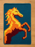 A knitted chunky blanket featuring a stylized horse rearing up. The horse is rendered in shades of orange, yellow, and maroon against a teal blue background. The piece has a textured, handmade appearance.