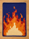 A chunky knit blanket featuring a vibrant flame pattern with cream, orange, and red stitches against a deep blue background, laid flat on a light wood floor.