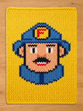 Chunky knit blanket in bright yellow featuring a pixelated-style knitted firefighter face with a blue helmet, mustache, and a red 'F' emblem, laid flat on a wooden surface.