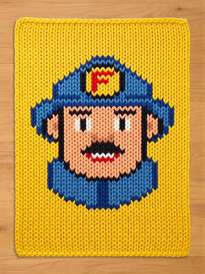 Chunky knit blanket in bright yellow featuring a pixelated-style knitted firefighter face with a blue helmet, mustache, and a red 'F' emblem, laid flat on a wooden surface.