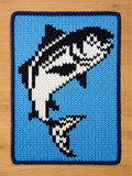 A knitted blue blanket with a navy blue border. The blanket features a black and white orca whale design in the center. The whale is jumping out of the water. The blanket is made of a soft, textured material.