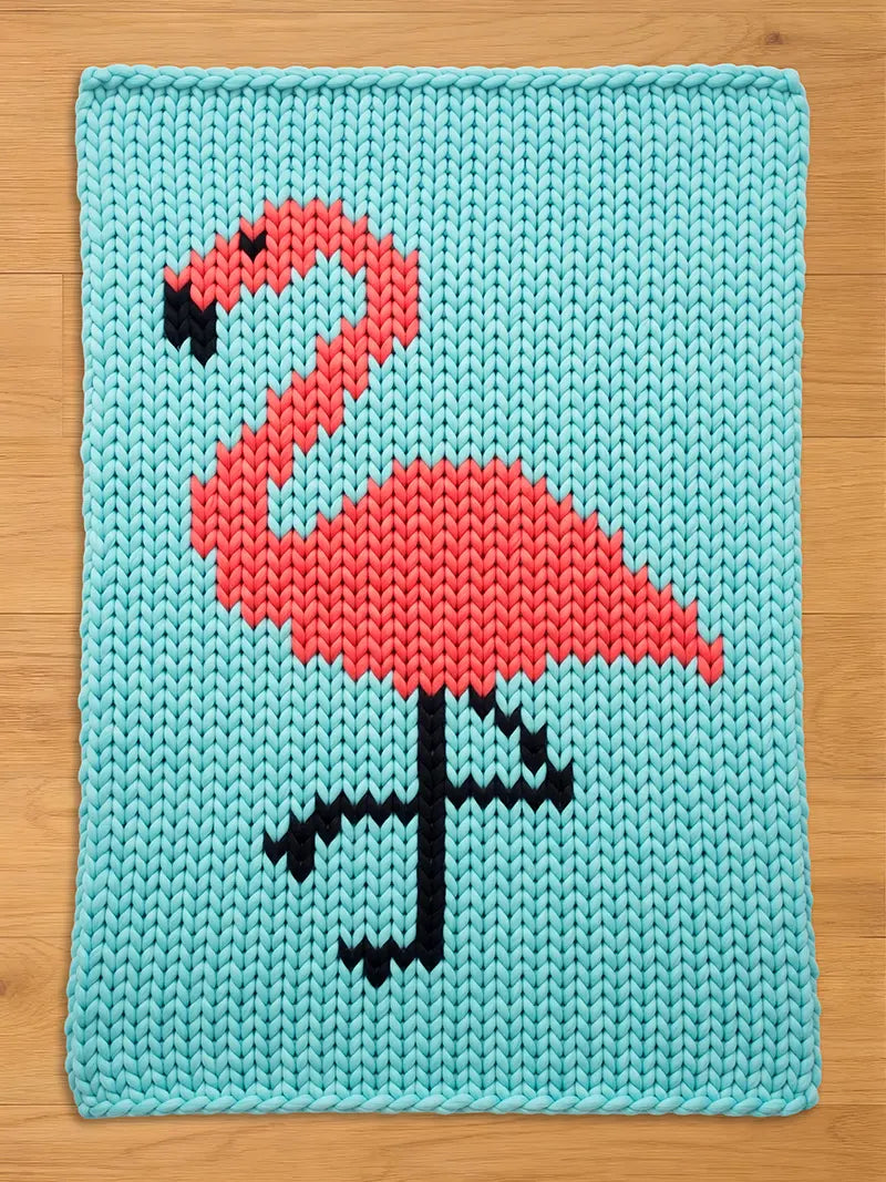 A knitted chunky blanket featuring a flamingo design. The flamingo is rendered in coral and black against a light blue background. The knitted texture is visible, adding depth to the design. The overall aesthetic is playful and decorative.