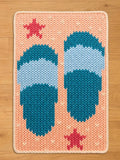 Knitted rectangular chunky blanket featuring a pair of blue and light blue sandals on a peach background. Two red star shapes are placed on the rug. The blanket has a cream-colored border.