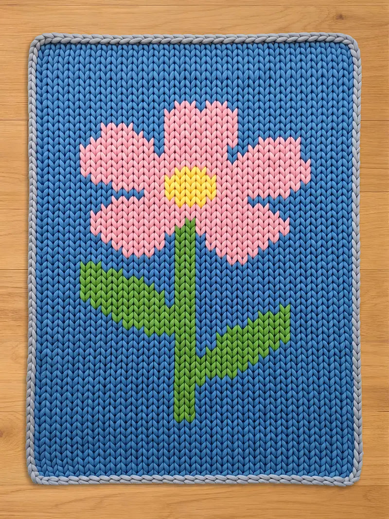Chunky knit blanket featuring a pink flower with a yellow center and green stem on a blue background, laid flat on a wooden surface.