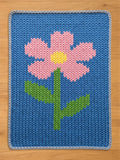 Chunky knit blanket featuring a pink flower with a yellow center and green stem on a blue background, laid flat on a wooden surface.