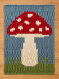 A rectangular knitted chanky blanket featuring a large red mushroom with white spots on its cap. The mushroom has a thick white stem and is positioned against a light blue background with green grass at the bottom edge of the piece.
