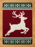 A chunky knit blanket featuring a large white silhouette of a leaping reindeer against a deep red background. The top and bottom borders have a green and white knit chevron pattern, displayed flat on a light wood floor.