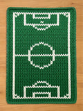 A green and white knitted chunky blanket featuring a soccer field design. The rug has a rectangular shape with a green background and white lines marking the field's boundaries, including the goal areas and center circle. The knitted texture adds a cozy, handmade feel.