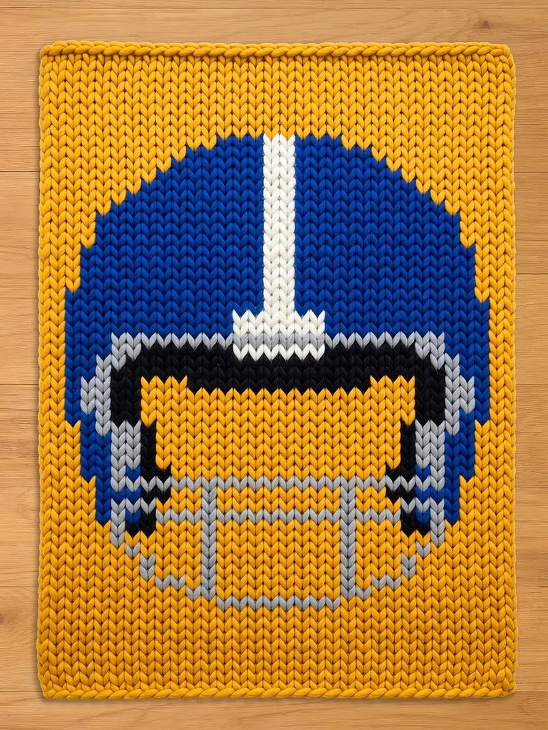A knitted throw blanket featuring a football helmet design. The helmet is blue with a white stripe, black, and gray details, set against a yellow background. The blanket is made of thick yarn.