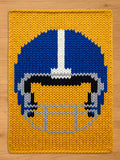 A knitted throw blanket featuring a football helmet design. The helmet is blue with a white stripe, black, and gray details, set against a yellow background. The blanket is made of thick yarn.